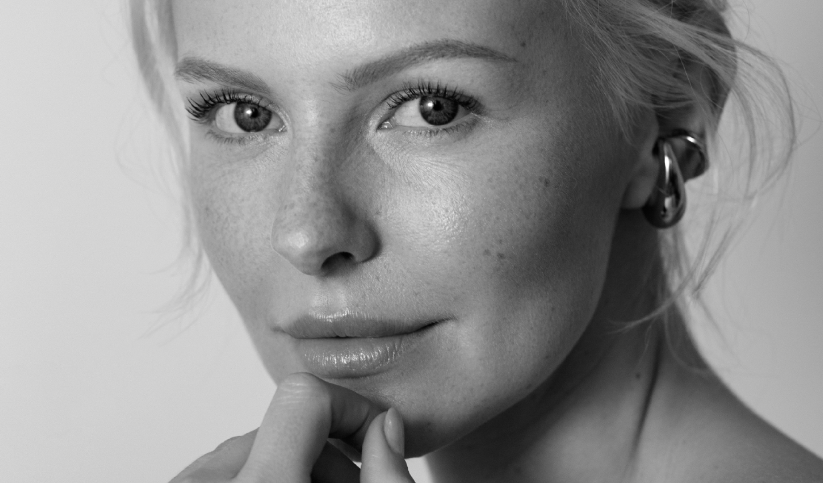 Black and white close-up portrait of a woman with light skin and light hair, looking at the camera with a gentle expression and resting her chin on her hand.